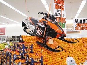 Hy-Vee employees hoisted a snowmobile atop a display of navel oranges. It was tethered  to support beams in the ceiling.