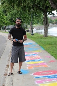 Artist Ryan Heath stands on his spray-on chalk artwork he painted on the Bridge Avenue sidewalk. The chalk washes away with rain. – Provided